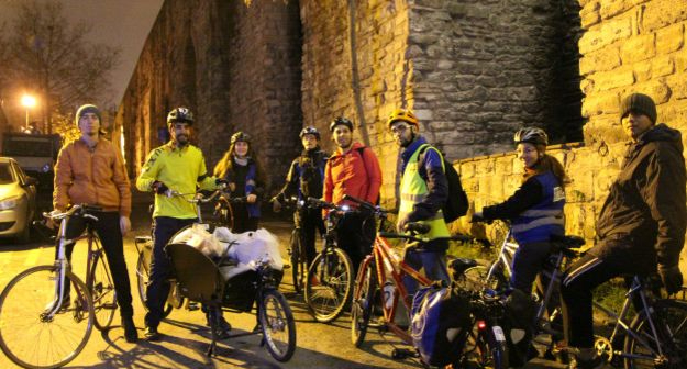 Gece geç saatlerde 1 kasalı bisiklet ve tandem bisikletlerle çorba dağıtımına çıkmış dernek gönüllüleri İstanbul surlarının yanında poz veriyorlar. Sokak ışıklarının vuran sarı rengi arka fon oluşturmuş.