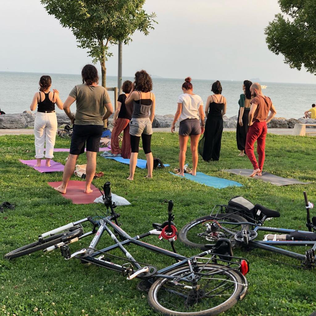 Çimlerin üzerinde matta 8 insan yüzleri denize dönük ayakta yogaya başlamaya hazırlanıyor. Hemen arkalaarında çimlere yatırılmış 2 adet tandem bisiklet bulunmaktadır.