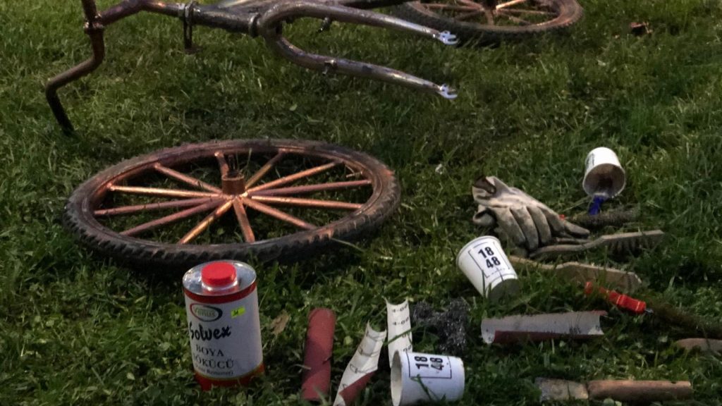 Çimlerin üzerinde zımpara ve boya için sökülmüş tekerlekler ve kadronun yanısıra boya sökücü, eldiven, zımbara, ve fırçalardan oluşan bakım ekipmanları bulunmaktadır.