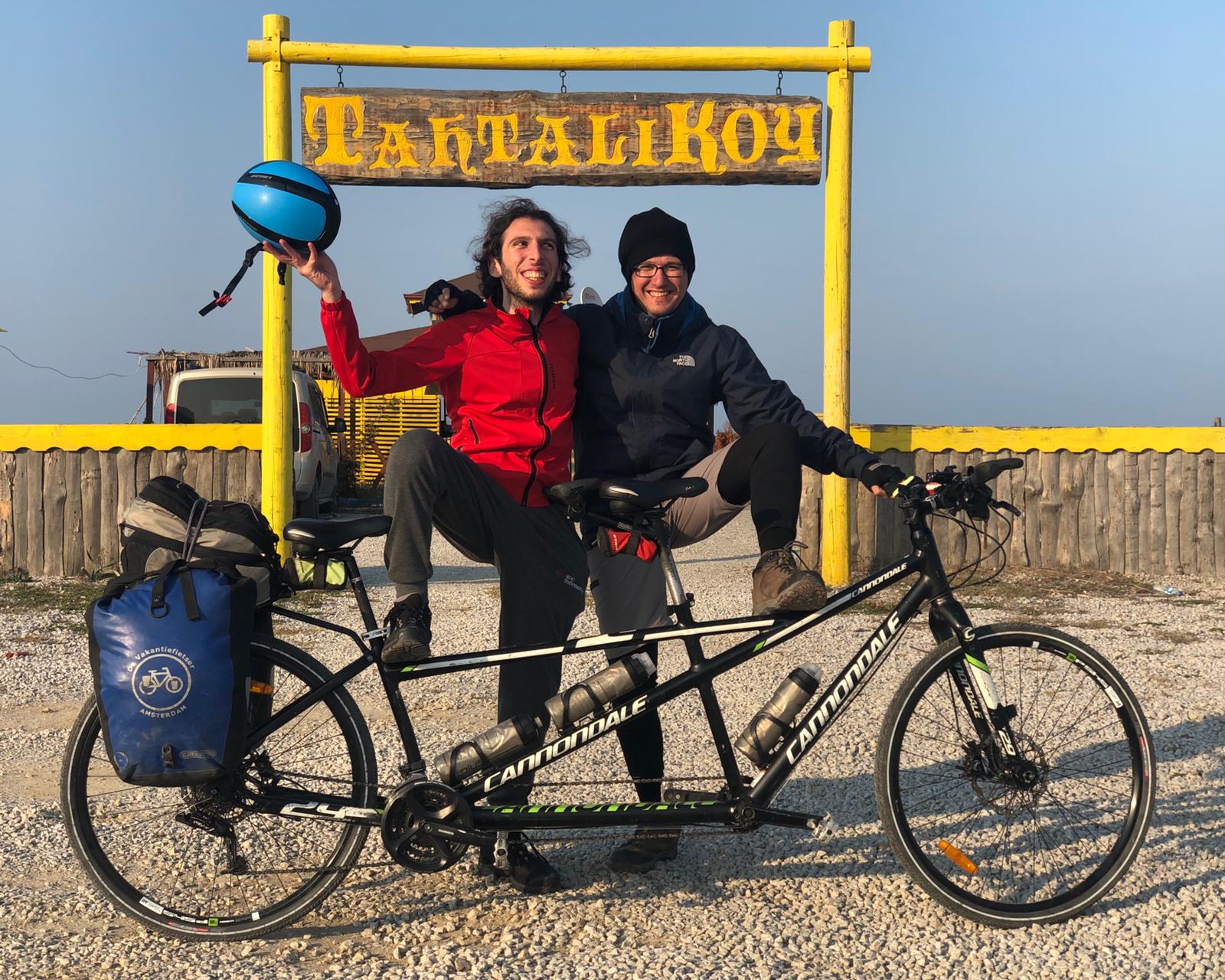 "Tahtalı Köy" tabelası önünde tandem bisikletle sağda Orhan Esen sol ayağını bisikletin ön selesine, Baran Altun ise sağ ayağını arka selesine v sağ elinde kaskı kaldırmış bir şekilde kameraya poz veriyorlar.